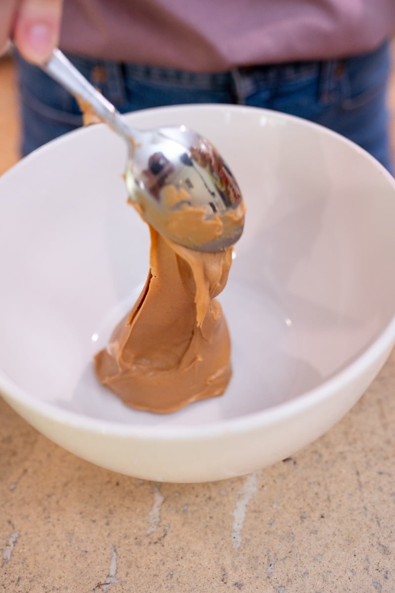 A spoonful of natural peanut butter dropping into a white mixing bowl