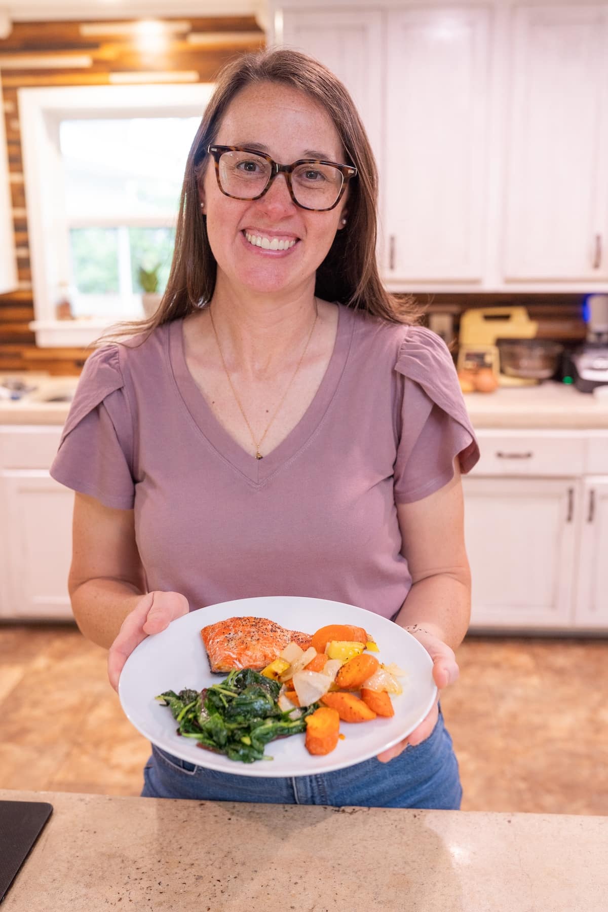 Sarah Bullard, MS,RD holding a balanced anti-inflammatory meal with salmon, vegetables, and greens in a home kitchen.