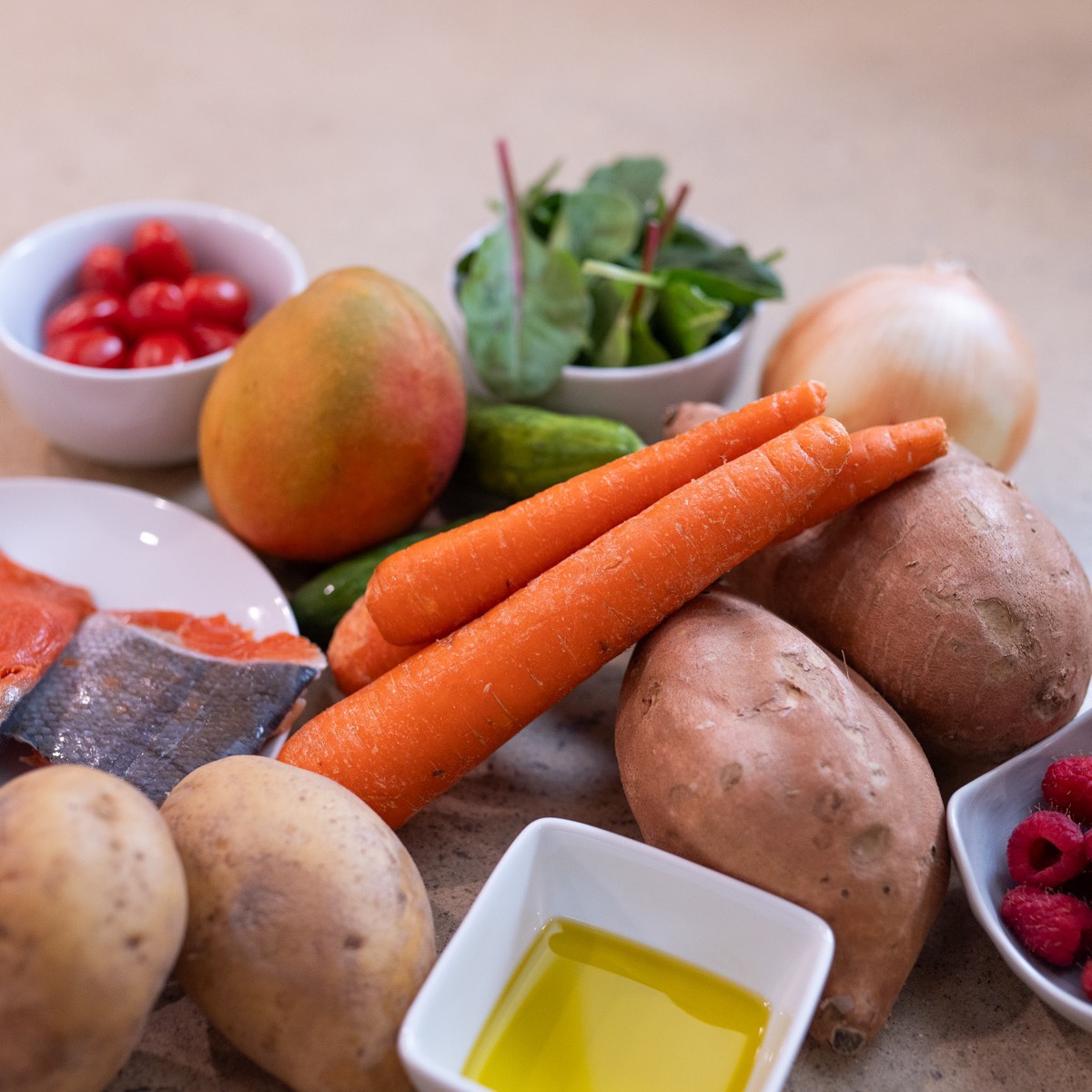 A spread of anti-inflammatory ingredients including carrots, cucumbers, leafy greens, sweet potatoes, berries and a dish of olive oil on a counter