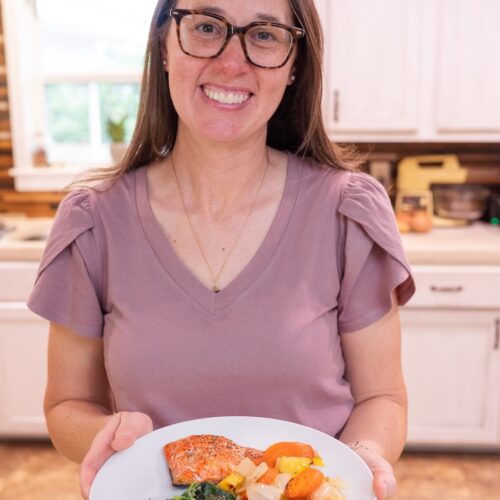 Sarah Bullard holding a white plate of baked salmon, roasted carrots and sweet potatoes, and sauteed leafy greens in her kitchen