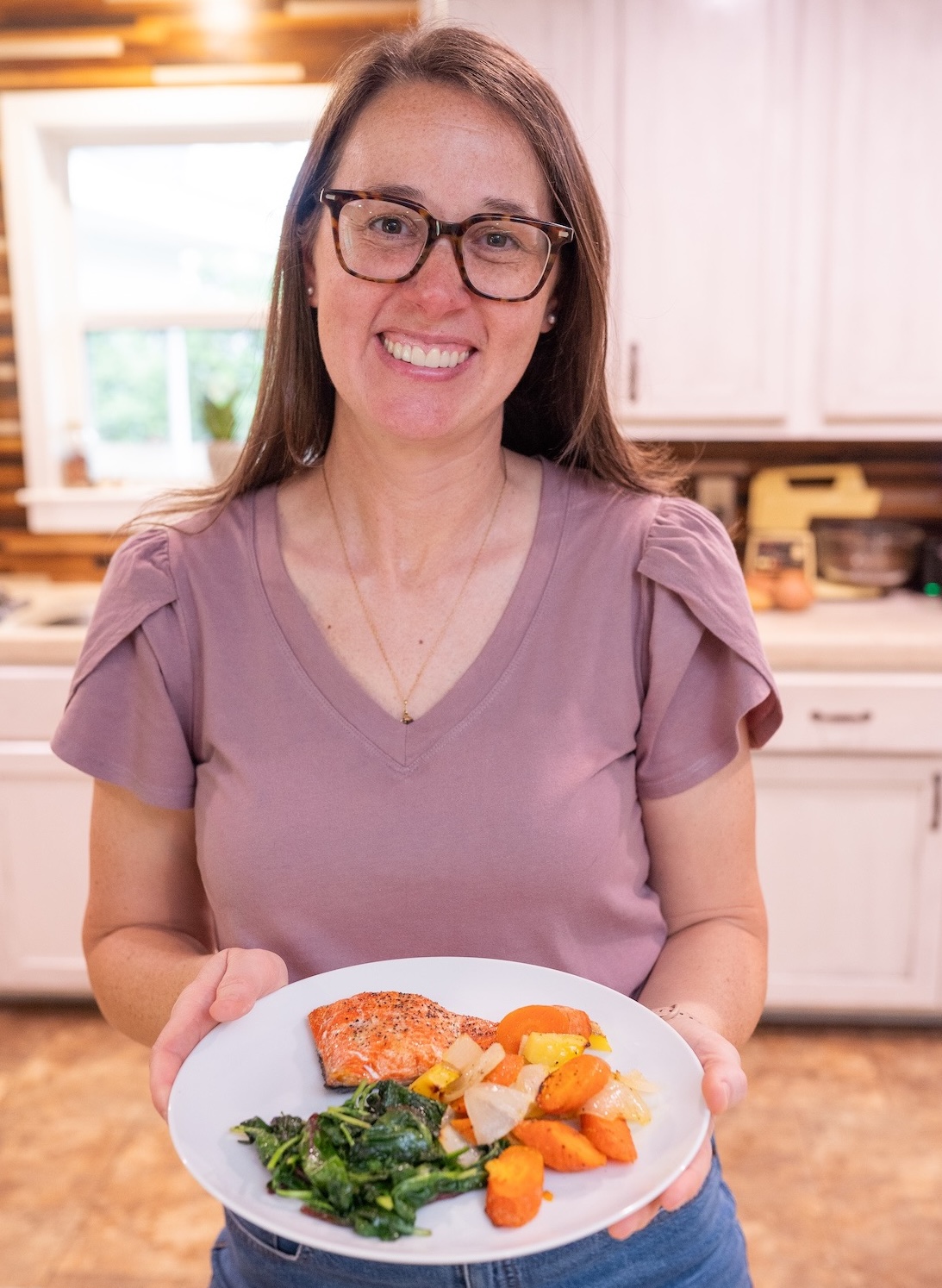Sarah Bullard holding a white plate of baked salmon, roasted carrots and sweet potatoes, and sauteed leafy greens in her kitchen