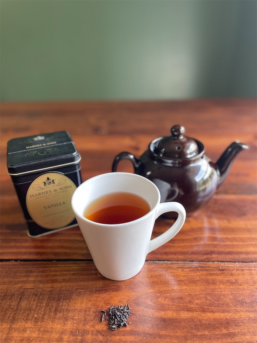 White mug of black tea with a brown teapot, a tin of Harney & Sons Vanilla black tea, and loose tea leaves on a wood table.