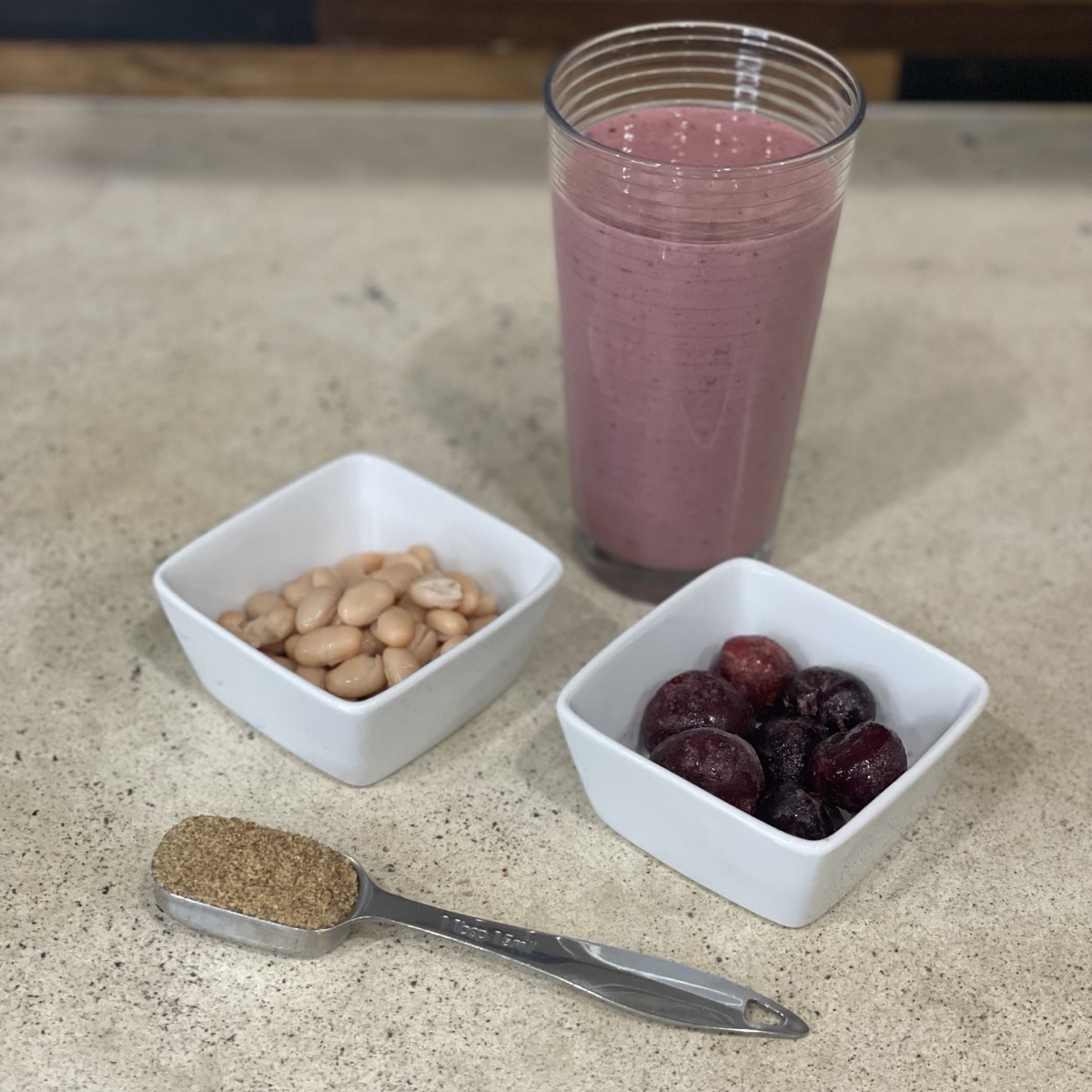 Pink cherry smoothie in a tall glass beside small bowls of white navy beans and frozen dark cherries, with a tablespoon of ground flaxseed