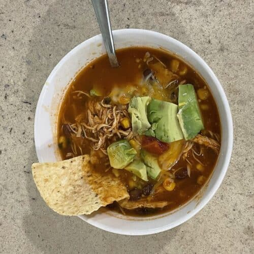 Overhead view of chicken tortilla soup in a white bowl, topped with diced avocado, shredded chicken, corn, black beans and melted cheese, with a tortilla chip resting on the rim.