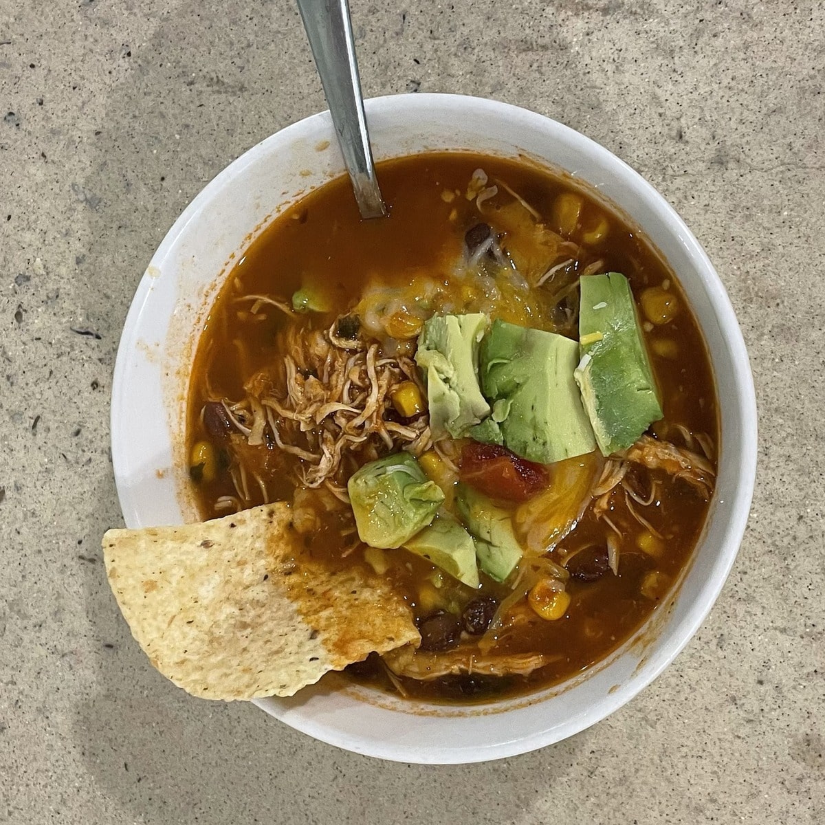 Overhead view of chicken tortilla soup in a white bowl, topped with diced avocado, shredded chicken, corn, black beans and melted cheese, with a tortilla chip resting on the rim.