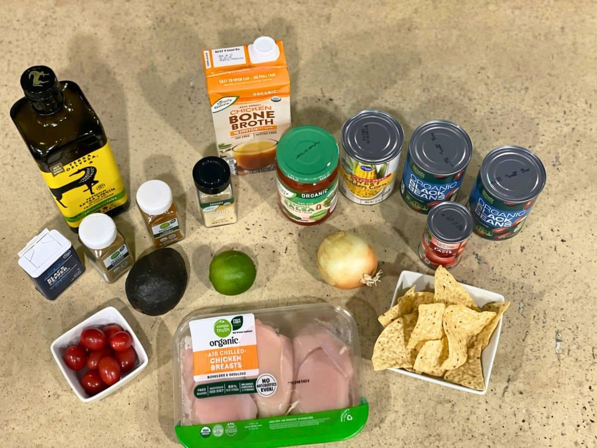 Ingredients for chicken tortilla soup laid out on a counter, including chicken breasts, bone broth, salsa, black beans, corn, tomato paste, onion, lime, avocado and tortilla chips.