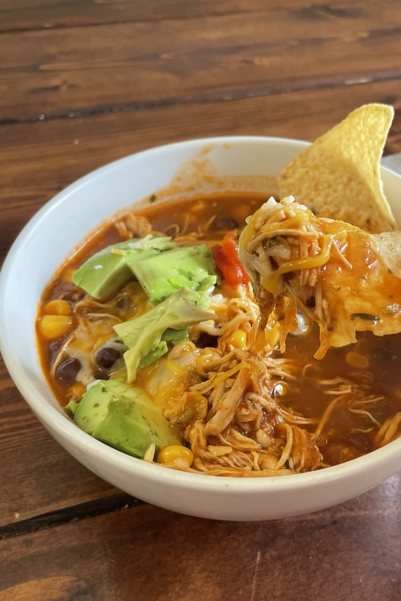 A tortilla chip lifted from a bowl of chicken tortilla soup, loaded with shredded chicken and melted cheese.