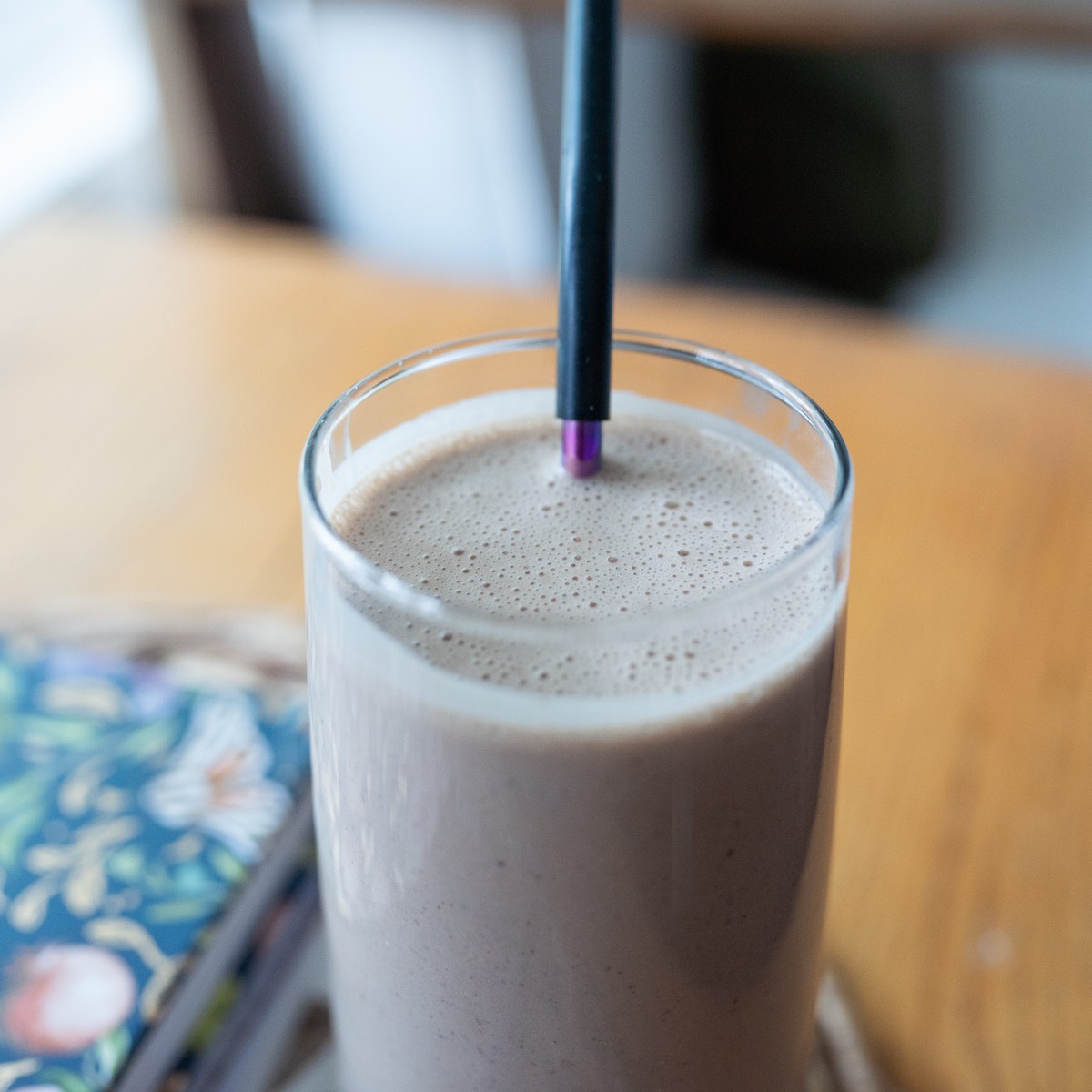 A chocolate banana smoothie in a tall glass with a black straw on a kitchen counter