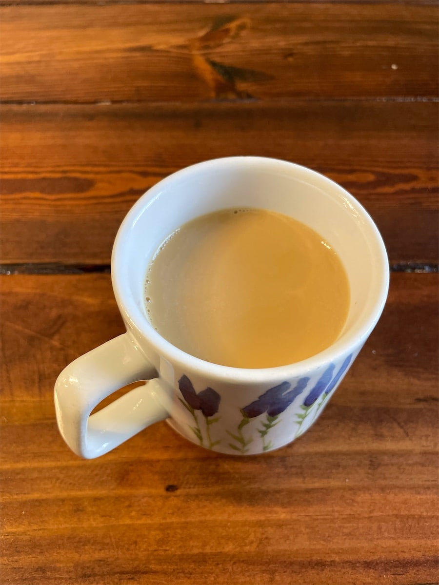Overhead view of coffee with half-and-half in a white mug with blue lavender flowers on a rustic wood table.