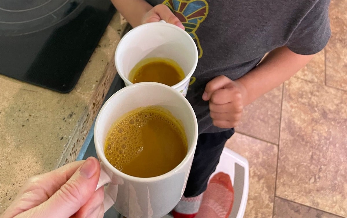 An adult's hand holding a white mug of golden milk next to a child's hand holding a smaller cup of golden milk by the kitchen counter.