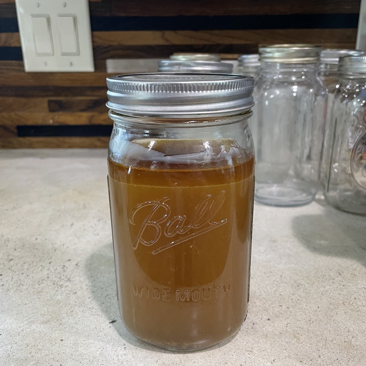 A wide-mouth Ball mason jar of golden beef bone broth on a kitchen counter, with empty jars and a wood plank wall behind it