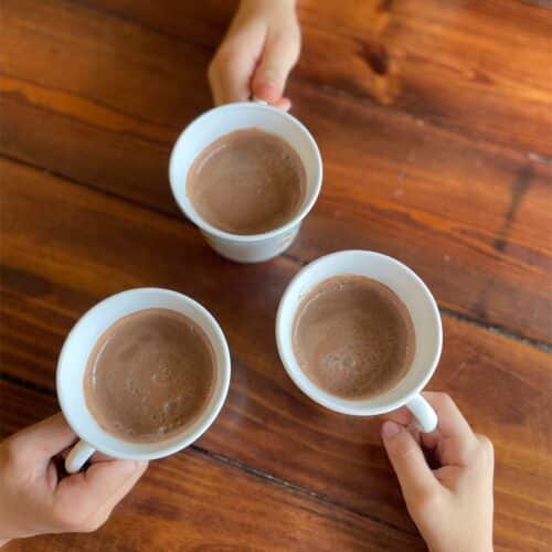 Three children's hands holding white mugs of hot cocoa, arranged in a circle on a wood table, viewed from above.
