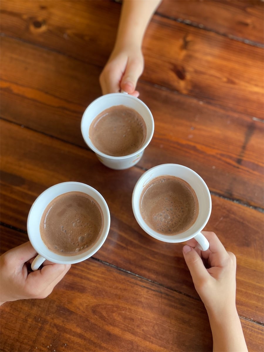 Three children's hands holding white mugs of hot cocoa, arranged in a circle on a wood table, viewed from above.