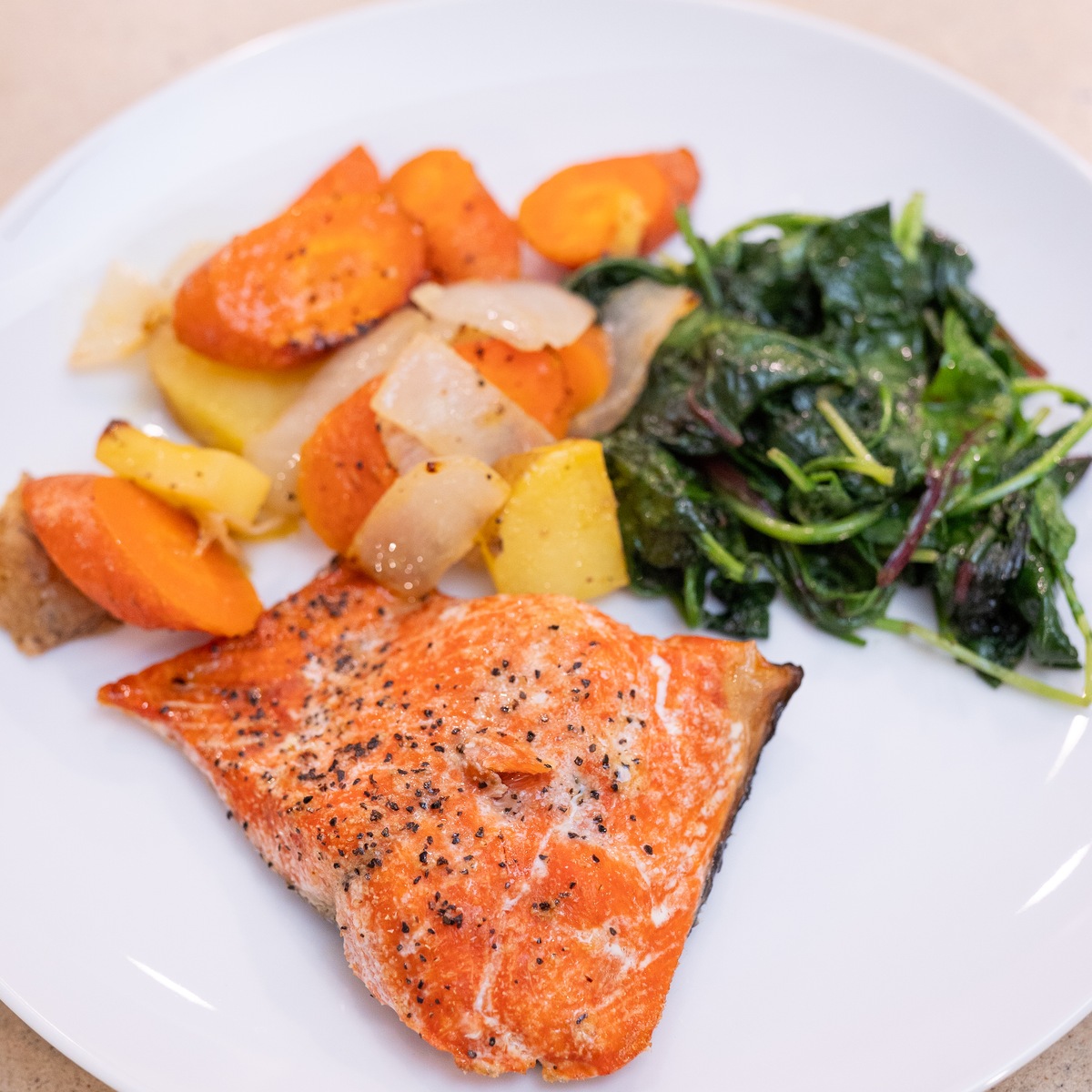 Baked salmon fillet on a white plate with sautéed greens, roasted carrots, potatoes and onion.