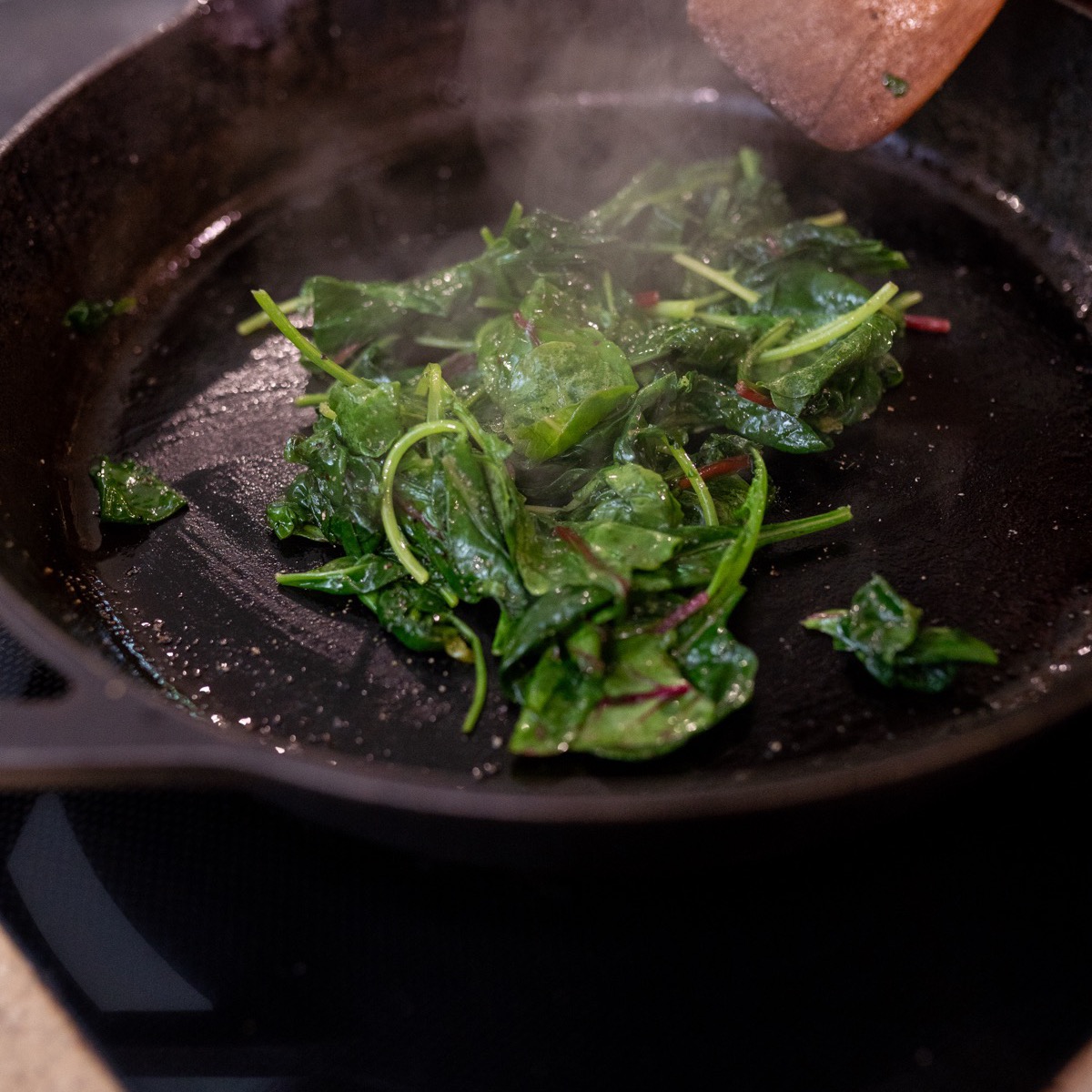 Leafy greens cooking in a cast iron skillet with steam rising and a wooden spoon