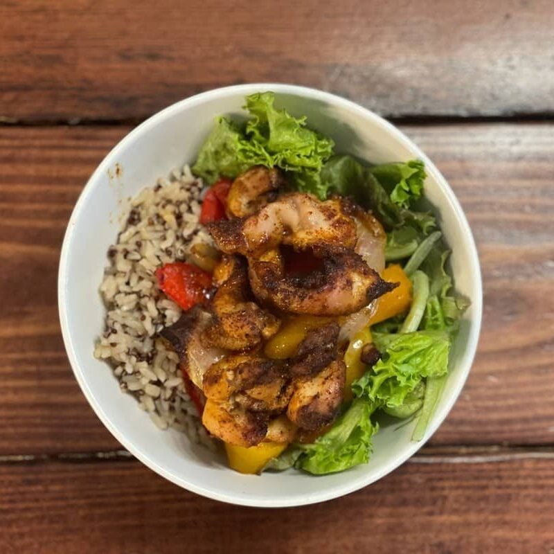 Overhead view of a chicken fajita bowl with roasted peppers, brown rice and quinoa, and leaf lettuce in a white bowl on a wood table