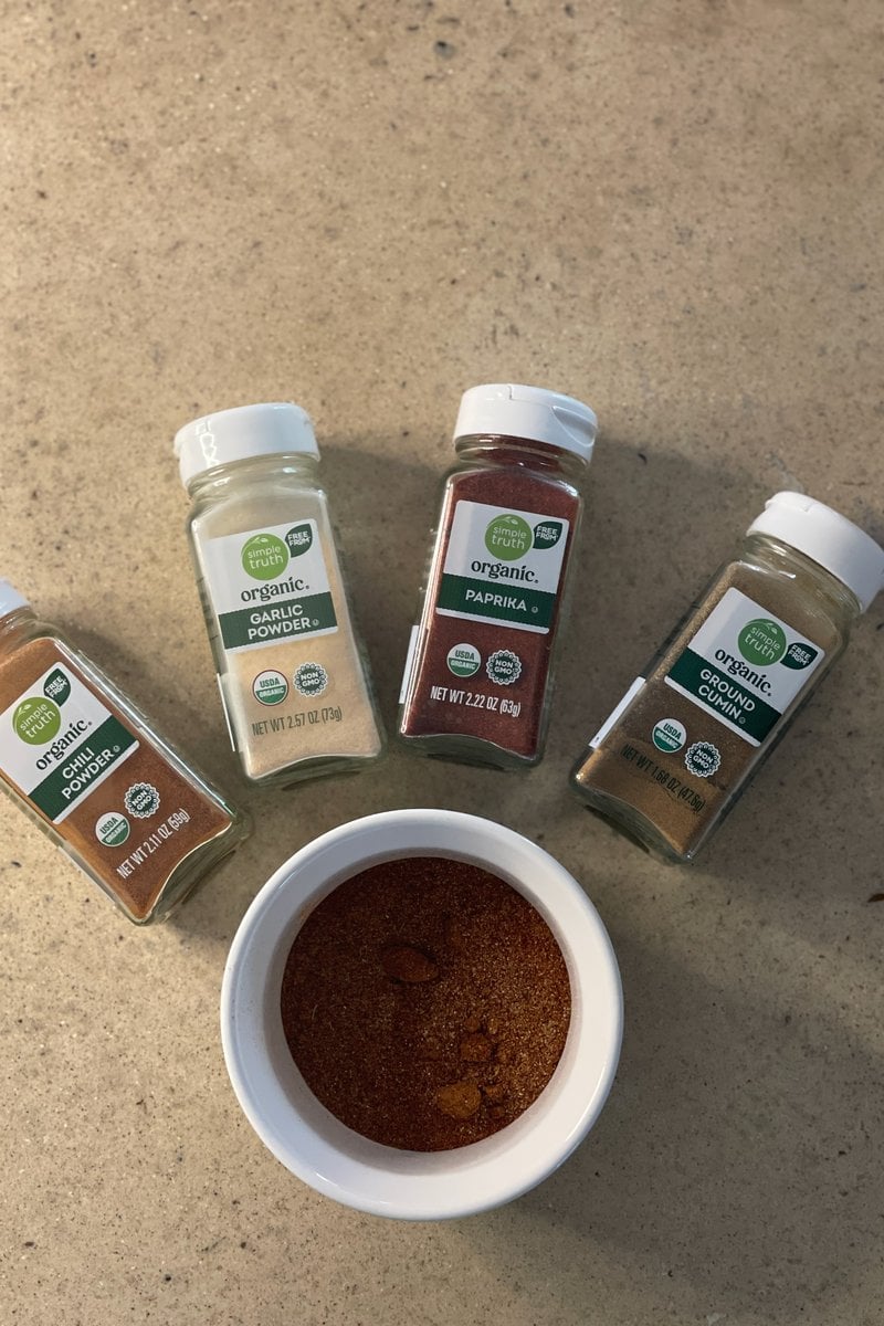 Chili powder, garlic powder, paprika and ground cumin jars around a small bowl of mixed fajita seasoning