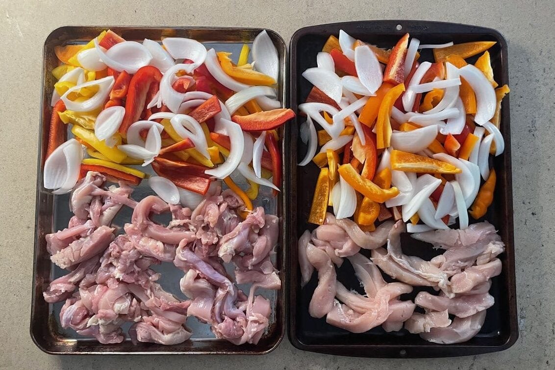 Two sheet pans of raw chicken thigh strips with sliced red, yellow and orange bell peppers and white onion