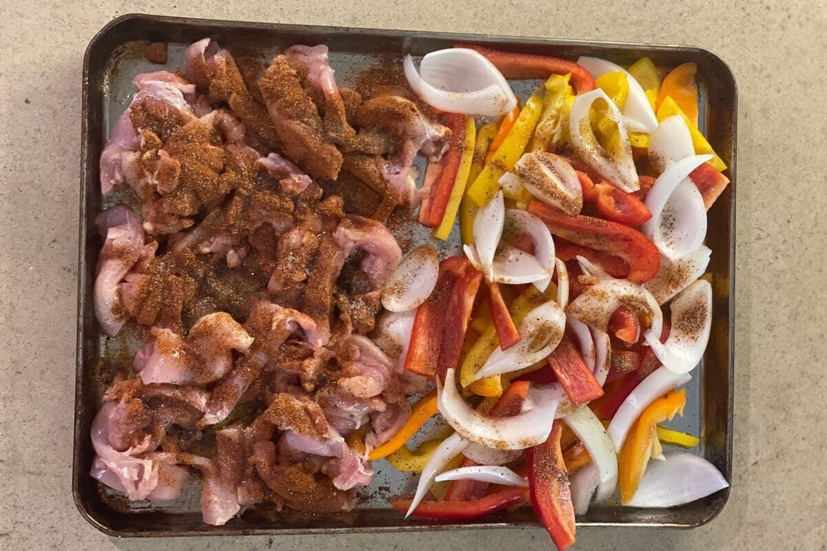 Sliced bell peppers, onion and raw chicken strips sprinkled with fajita spices on a sheet pan, ready for the oven