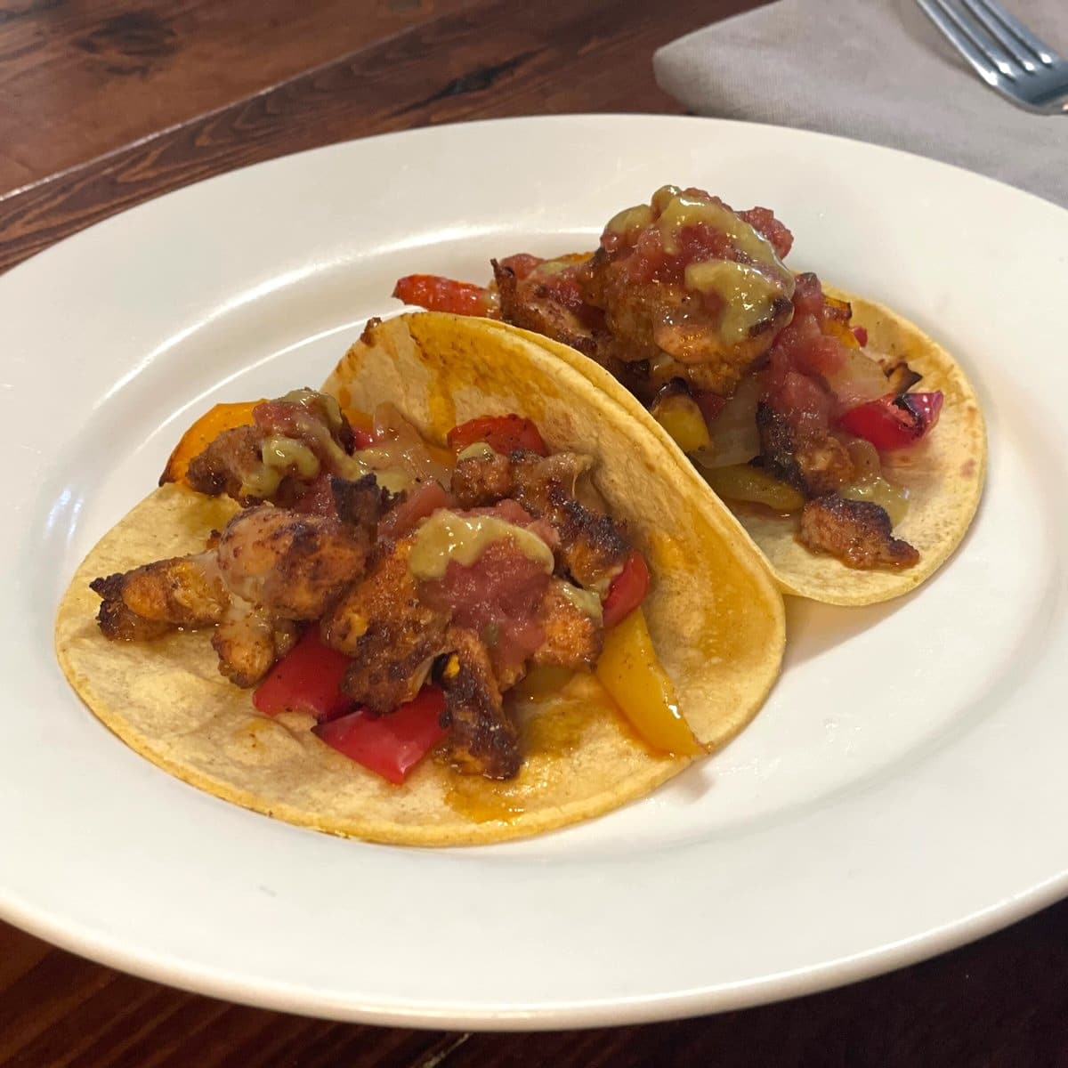 Two sheet pan chicken fajita tacos in corn tortillas with roasted red and yellow peppers, salsa and a green jalapeño sauce on a white plate